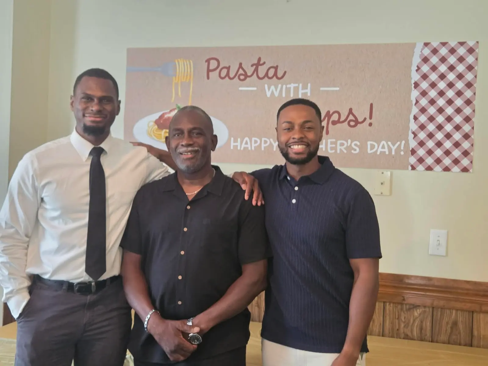 Three men smiling in front of the Pasta with Pops Father's Day banner