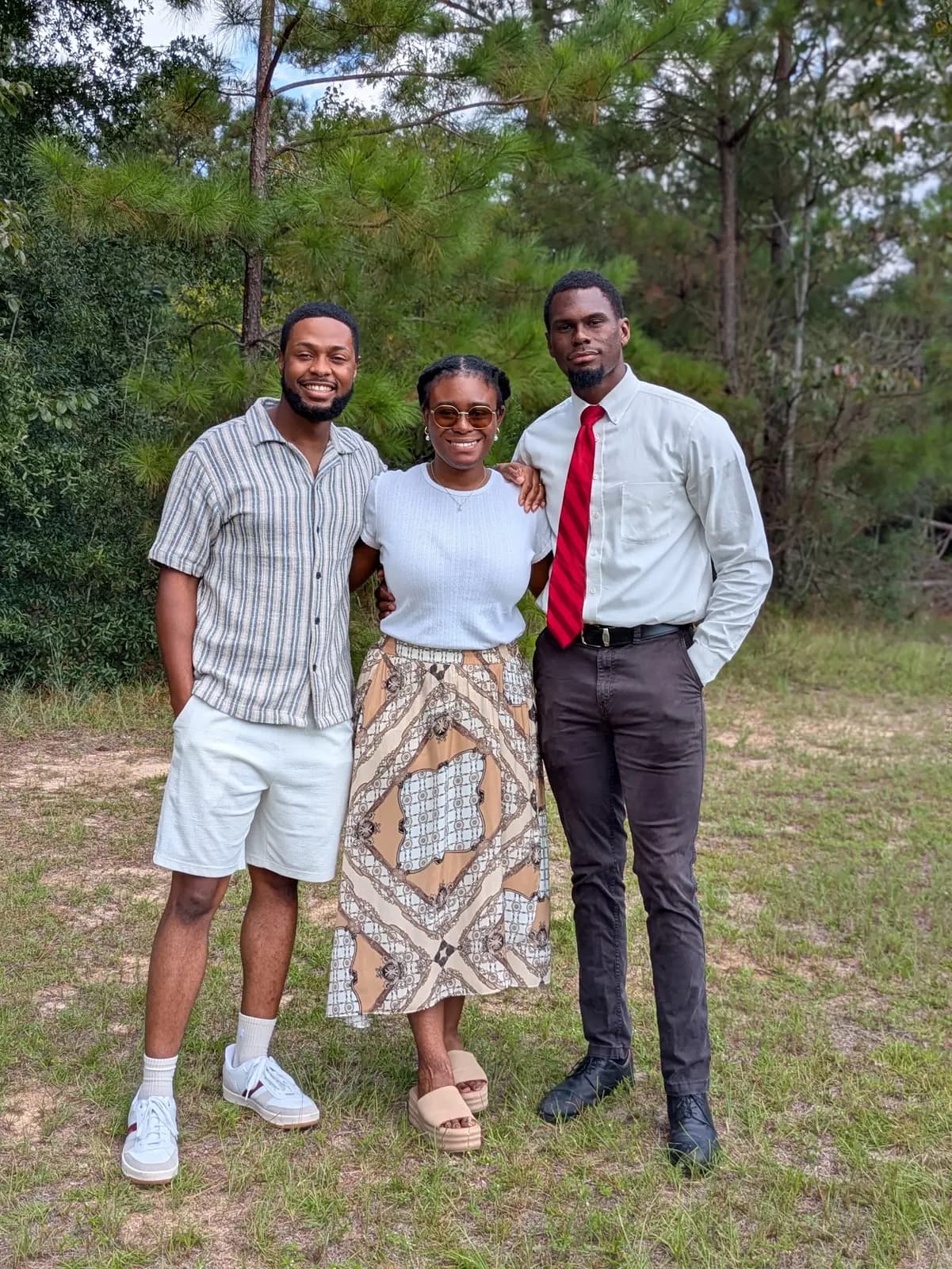 Three friends smiling outdoors in front of tall pines